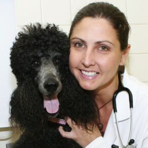 A photo of Dr. Deirdre Chiaramonte with her white coat and stethoscope and her black poodle heart dog, Dijon, AKA Poopie.